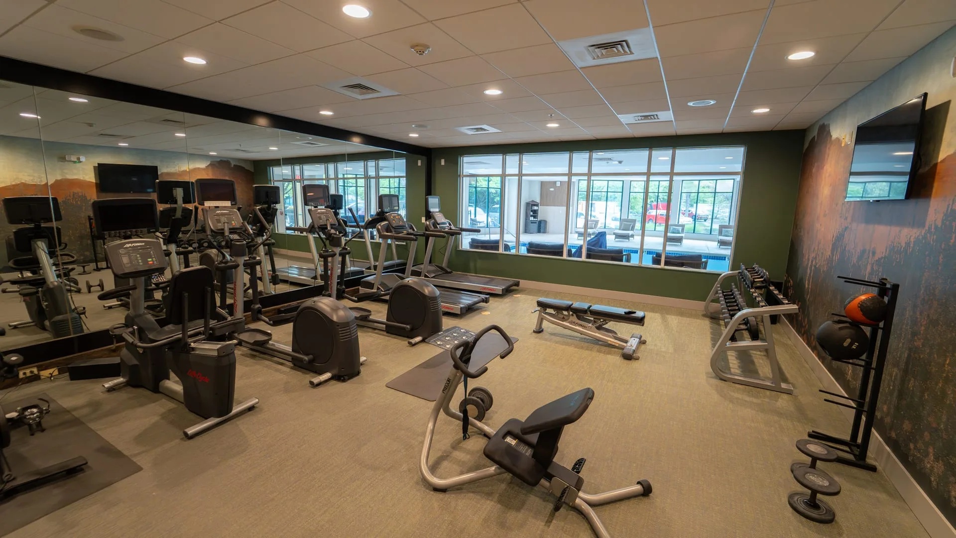 Hotel fitness center with mirrored walls, exercise equipment and windows overlooking the indoor pool